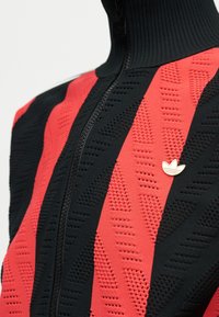 Lähivõte musta ja punase kootud jaki kõrge krae, esiosa tõmbluku, tekstuurse mustri ja väikese valge Adidas trefoil logoga rinnal.