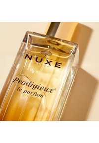 Klart glasparfymflaska med en gyllene vätska. Har ett guldlock och svart text. Logotyp och produktnamn synliga framifrån.