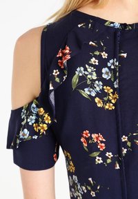 Sukienka w kwiaty w kolorze granatowym, z krótkimi rękawami, falbanym wykończeniem i zapięciem na guziki. Wzór zawiera niebieskie, żółte i pomarańczowe kwiaty.