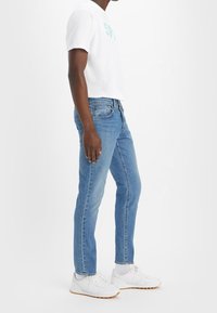 T-shirt blanc à manches courtes avec un graphisme bleu sarcelle, associé à un jean en denim bleu clair et des baskets blanches. Coupe droite, style décontracté.