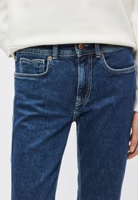 Vaqueros de mezclilla azul oscuro con corte ajustado, que presentan detalles de costura, cinco bolsillos y un cierre con botón de cobre en la cintura.