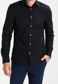 Camicia nera a maniche lunghe con colletto a camicia, bottoni bianchi e finitura in tessuto liscio. Vestibilità sartoriale e design minimale.