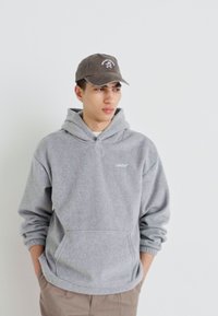 Felicetta grigia con cappuccio e tasca frontale, caratterizzata da un piccolo logo bianco. Abbinata a un cappello grigio scuro con dettagli ricamati.