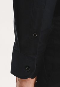 Czarny bawełniany mankiet koszuli z dwiema czarnymi guzikami, charakteryzujący się gładką strukturą i dopasowanym krojem. Mankiet lekko wystaje ponad nadgarstek.