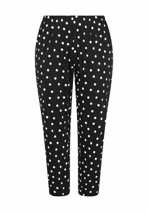 Donna con lunghi capelli biondi che indossa una maglietta nera "Sleepover Club" e pantaloni neri con pois bianchi, in piedi davanti a uno sfondo bianco.