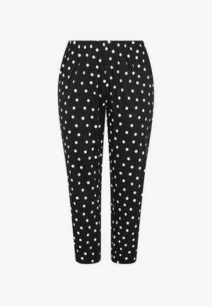 Vrouw met lang blond haar die een zwart "Sleepover Club" T-shirt en een zwarte broek met witte stippen draagt, staande tegen een witte achtergrond.