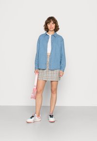 Camisa de ganga, top corto branco, saia mini xadrez, bolsa de ombro rosa e sapatilhas brancas com detalhes em rosa. Modelo de pé contra um fundo simples.