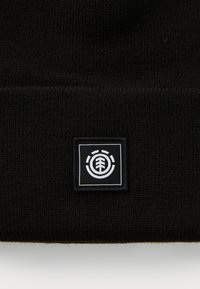 Bonnet en tricot noir avec un revers, arborant un écusson carré en noir et blanc représentant un design d'arbre. Tissu doux et texturé.