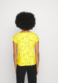 Person med lockigt hår som bär en kortärmad gul skjorta med ett brunt kedjemönster och svarta byxor, sedd bakifrån mot en vit bakgrund.