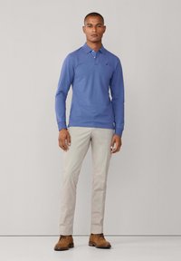 Dlhý rukáv polokošeľa z ľahkej modrej bavlny, kombinovaná s béžovými nohavicami slim-fit a hnedými šnurovacími topánkami, stojaca pred neutrálnym pozadím.