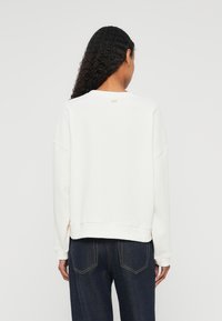 Sweatshirt branca com decote arredondado e ombros caídos, apresentando um acabamento texturizado e um pequeno logo nas costas. Combinado com denim escuro.
