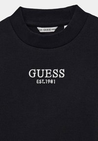 Pull noir côtelé avec un col haut et le logo "GUESS EST. 1981" brodé en blanc sur le devant. Texture douce avec une coupe ajustée.