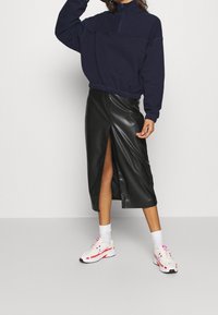 Sweat-shirt marine avec un col à fermeture éclair quart de tour, associé à une jupe midi en simili cuir noire avec une fente à l'avant, et des baskets blanches avec des accents roses.