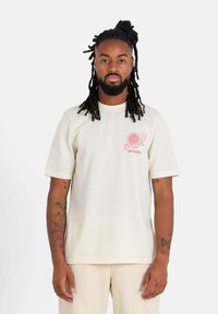 Crème korte mouwen t-shirt met een roze bloemenafbeelding op de linkerkant van de borst, gemaakt van zacht katoen, met een relaxte pasvorm en een ronde hals.