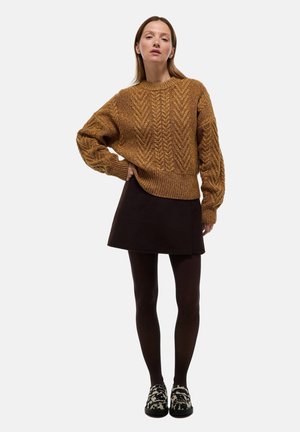 Pull en maille torsadée marron avec un ourlet côtelé, associé à une jupe sombre, des collants noirs et des chaussures à enfiler à motifs. Fond de studio.