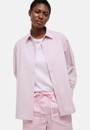 Jeune femme portant une chemise à rayures roses claires boutonnée sur un t-shirt blanc et un pantalon à cordon rose assorti, debout devant un fond blanc.