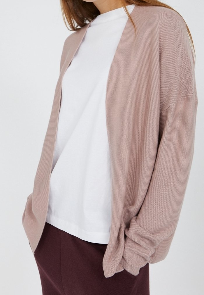 Světlá růžová kardigan z texturovaného pleteného materiálu se volným střihem a otevřeným předním dílem, nošený přes bílé tričko a vínově červené kalhoty.