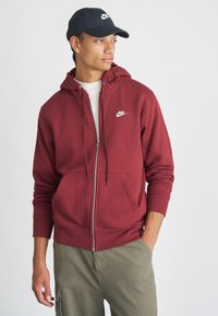 Sweatshirt marrom de fecho com capuz, feito de tecido macio, com um bolso frontal e capuz ajustável com cordão; apresenta um logo branco da Nike no peito.