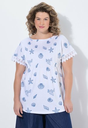 Kobieta o kręconych włosach nosi białą bluzkę z niebieskimi nadrukami o tematyce morskiej i rękawami wykończonymi koronką, do tego ciemnoniebieskie spodnie, na białym tle.