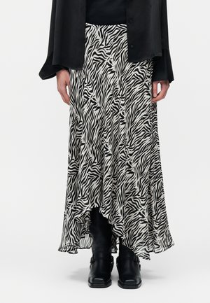 Frau trägt einen schwarz-weißen Rock mit Zebra-Muster, der vorne kurz und hinten lang ist, dazu ein schwarzes Oberteil und schwarze Stiefeletten, stehend vor weißem Hintergrund.