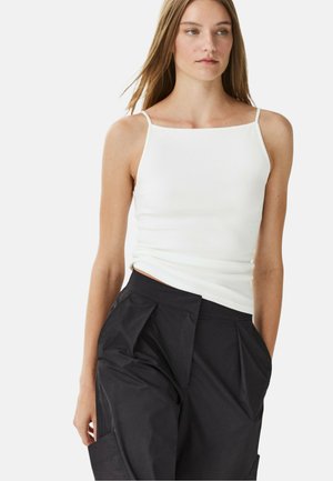 Giovane donna che indossa un top bianco senza maniche con spalline sottili e pantaloni neri a vita alta con tasche, in piedi davanti a uno sfondo semplice.