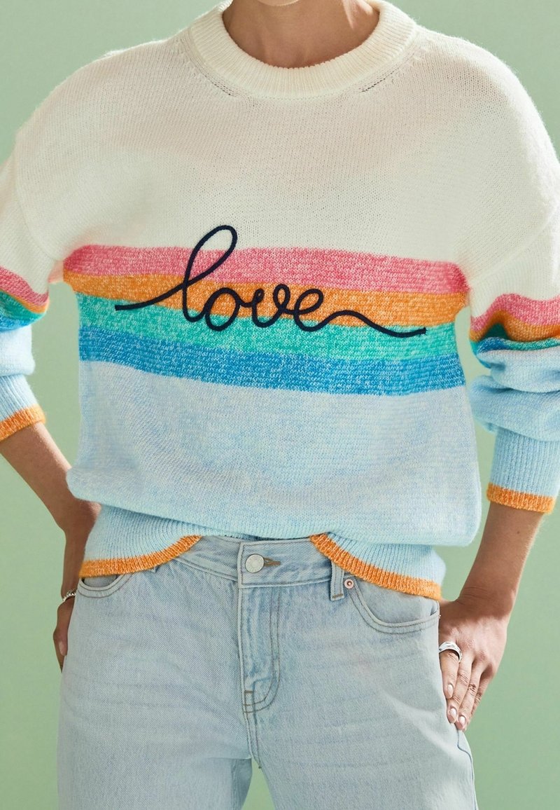 Pull avec des rayures horizontales aux couleurs de l'arc-en-ciel et un texte noir en cursive « love » sur la poitrine, associé à un jean bleu clair et les mains sur les hanches.