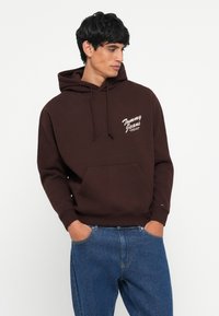 Bruine hoodie met een voorzak, trekkoorden en wit "Tommy Jeans" logo. Gemaakt in combinatie met blauwe spijkerbroeken.