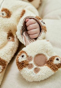 Un indumento in pile color crema presenta maniche imbottite con design di zampe di orso. All'interno ha strisce alternate marroni e crema. Si nota una texture morbida.