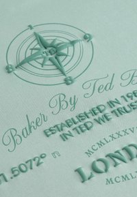 Tekstūrēts piparmētru zaļš papīrs ar reljefiem kompassa dizainiem, stilizēta teksta "Baker By Ted Baker" un koordinātām, kas izceļ sarežģītas detaļas.