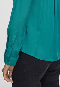 Blusa di raso verde acqua con una texture liscia, polsini con bottoni e orlo curvo, abbinata a pantaloni neri con una tasca posteriore.