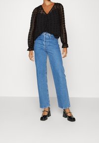 Camicia nera trasparente con pois testurizzati, scollo a V profondo e maniche lunghe. Abbinata a jeans in denim blu chiaro a gamba dritta e scarpe con tacco nere.
