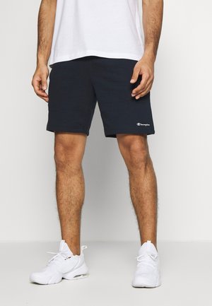 Mand iført mørkeblå Champion-shorts, hvide sneakers og en hvid t-shirt, stående mod en ensartet lys baggrund.