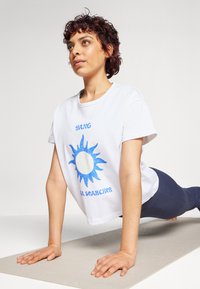 Bijela pamučna majica s plavim grafičkim dizajnom sunca i natpisom "YANG YOGA SEARCHER." Uparena s tamnoplavim tajicama, model u položaju daske.