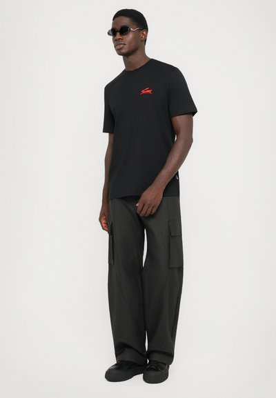 Sort bomulds T-shirt med rødt logo, parret med løstsiddende sorte cargo bukser med sidelommer, og sorte slip-on sko.