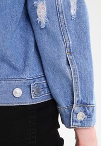 Ljust blå denimjacka med slitna partier, silverfärgade knappstängningar i ärmslut och orange sömmar längs sömmarna.