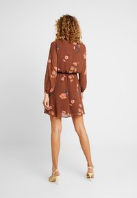 Brun blommig klänning med långa ärmar, åtdragen midja och genomskinligt tyg. Har rosa och blå blommönster på en enfärgad bakgrund.