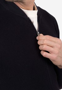 Suéter azul marino con textura y cremallera, con cuello simulado, mostrando una mano cerrando la cremallera y una camisa blanca debajo. Cuenta con herrajes de cremallera metálicos.