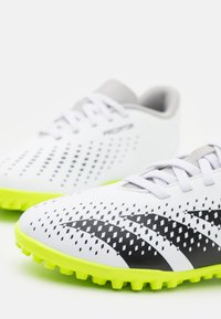 Scarpe da calcio bianche con superficie testurizzata, strisce nere, suole giallo-verdi e accenti grigi; design traspirante con perforazioni.