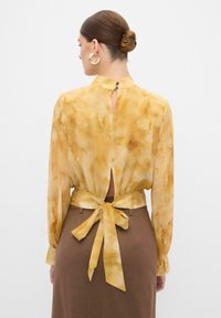 Gelbe Bluse mit einer Binde-Schlaufe, die ein Schlüsselloch-Öffnung am Nacken hat. Der Stoff hat eine weiche, fließende Textur mit subtilen marmorierten Mustern.