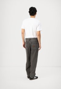 Grijze denim broek met een losse pasvorm, voorzien van achterzakken en een kleurrijke logopatch. Draagt een eenvoudig wit T-shirt en zwarte schoenen.