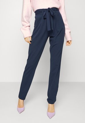 Femme portant un pantalon paperbag bleu marine taille haute avec une ceinture nouée, associé à un pull rose clair et des talons pointus roses.