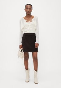 Un cardigan court à manches longues blanc sur un haut crème avec de petits points, associé à une mini jupe en daim marron et des bottes de cowboy blanches.