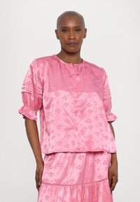Rosa satengbluse med blomsterdesign, rund halsutskjæring, knappelukking og korte, volang-ermer. Teksturen ser glatt og blank ut.