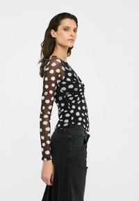 Top a maniche lunghe in tessuto nero trasparente con pois bianchi, caratterizzato da una silhouette aderente e un collo rotondo. Indossato con jeans scuri.