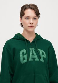 Vihreä vetoketjuhuppari, jossa on huppu ja erottuva vihreä "GAP" -logo. Huppari on valmistettu pehmeästä puuvillamateriaalista ja siinä on etupuolella kengurupockets.
