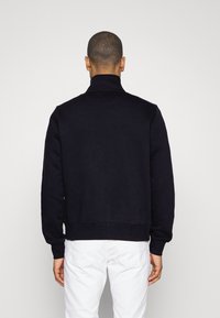 Felpa blu navy con colletto alto, polsini e orlo a costine. La vista posteriore mostra una texture liscia e un design minimalista, caratterizzato da una sottile cucitura a V.