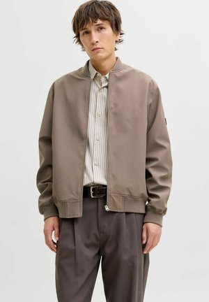 Ung mand iført beige bomberjakke, stribet skjorte med knapper og brune bukser, stående mod en ensfarvet lys baggrund.