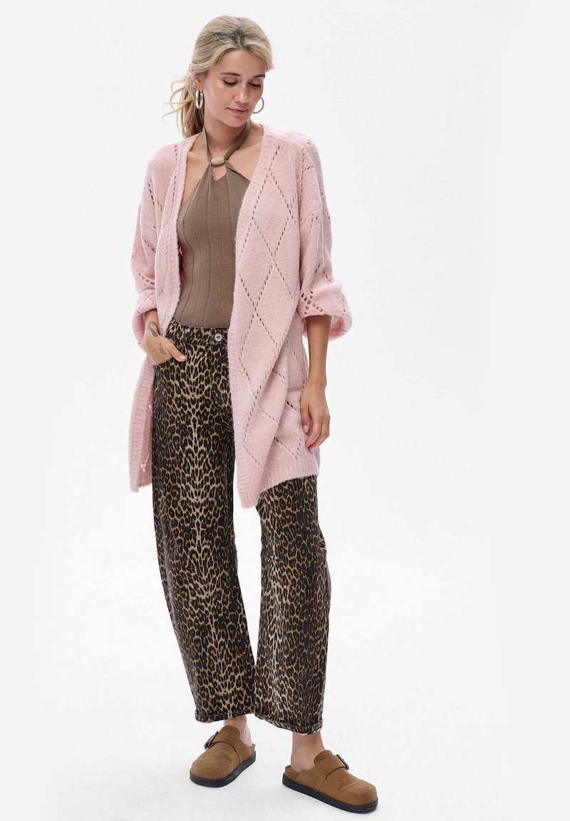 Lichtroze gebreide cardigan met diamantpatroon, gedragen over een bruine top en brede broek met dierenprint. Bruine instappers maken de outfit compleet.