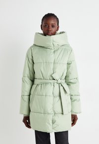 Manteau matelassé en vert clair avec un col haut, une taille ceinturée et des détails de couture visibles. Texture douce et coupe ample.