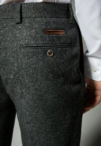 Pantalones grises texturizados que cuentan con un bolsillo trasero con un acento de cuero marrón y un cierre de botón. El material tiene un aspecto parecido a la lana con un patrón moteado.